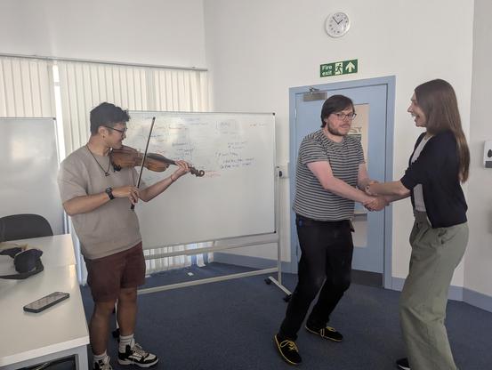 Dr Andrew Spencer and Dr Rachel Grey practising the steps with Lewis Hou of Science Ceilidh on the fiddle. They are in font of a white board relating how the inspiral, merger and ringdown phases of a binary black hole coalescence can be turned into a dance.