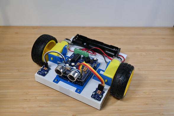 Front view of a simple robot constructed on a white wooden base. The robot is controlled by an Arduino UNO with an ARPS-2 shield mounted on top. A SONAR module is plugged into ARPS-2, and two optical floor sensor modules have been snapped off ARPS-2, mounted on the wooden base, and connected to ARPS-2 using short extension cables. The robot is propelled by two inexpensive yellow gear motors connected to yellow and black wheels, and powered by AA batteries (not shown) that would be installed in a battery holder at the back.