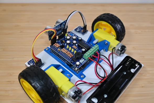 Rear side view of a simple robot constructed on a white wooden base. The robot is controlled by an Arduino UNO with an ARPS-2 shield mounted on top. A SONAR module is plugged into ARPS-2, and two optical floor sensor modules have been snapped off ARPS-2, mounted on the wooden base, and connected to ARPS-2 using short extension cables. The robot is propelled by two inexpensive yellow gear motors connected to yellow and black wheels, and powered by AA batteries (not shown) that would be installed the black battery holder. The motors and Arduino are mounted to the base using blue, 3D-printed mounts.