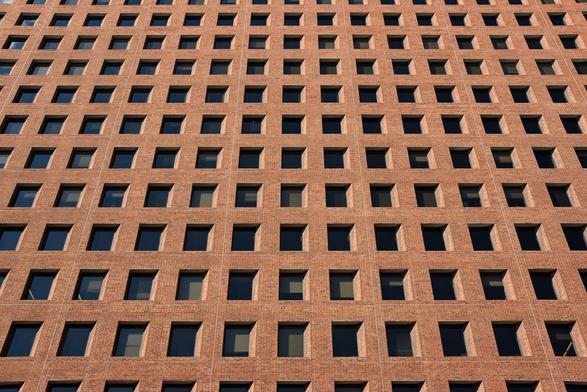 Color photo looking up at the façade of a brick clad office building. Regularly spaced square windows - each of which displays a deep, angled reveal - give the wall the appearance of an extensive grid.