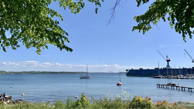 Das Bild zeigt eine ruhige und idyllische Küstenlandschaft. Im Vordergrund sind grüne Bäume zu sehen, die einen natürlichen Rahmen bilden. Der Hintergrund besteht aus einem ruhigen, blauen Wasser, auf dem ein Segelboot und ein kleinerer roter Bootsschiffchen schwimmen. Auf der rechten Seite des Bildes befindet sich ein Hafen mit mehreren Schlepper und Kränen, die auf Schiffsbau oder Reparaturen hinweisen. Ein Holzsteig führt ins Wasser und bietet Zugang zum Hafen. Im Hintergrund sind sanfte Hügel und ein klarer, blauer Himmel mit einigen weißen Wolken zu sehen. Die Szene wirkt friedlich und einladend, mit einer Mischung aus Natur und menschlicher Aktivität.