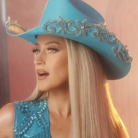 A blonde woman with pin straight hair and pale blue eyes looks off to the right. She wears a powder blue cowboy bat and sleeveless vest with a floral rhinestone design.