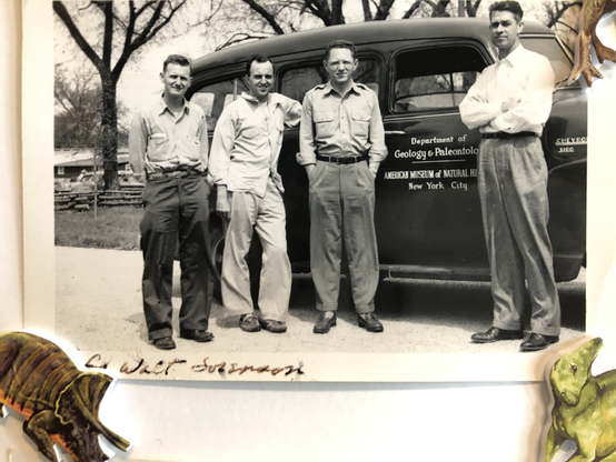 Black and white photo of four men in front of a large 1940s era Chevrolet Suburban. They appear to be wearing khakis and similar outfits of the style popular in the 1940s.