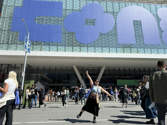 Chi posing with her arms and one leg raised in front of Moscone Center on a nice sunny day, with the first 3 stylized letters or glyphs of "Config" shown above her on the glass windows of the building. Lots of other conference goers are seen walking around and about behind Chi.