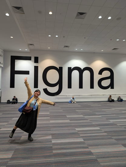 Chi posing with her arms and one leg raised gesturing to the huge "Figma" sign stuck on the white wall behind her in the tall hallway of Moscone Center. Ceiling lights shine spotlights on the wordmark and Chi's position puts her under the i of the logo from the perspective of the image. Other conference goers are also visible nearer the wall.