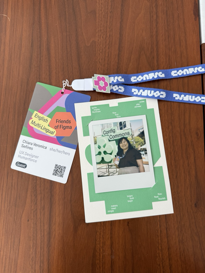 Flatlay of Chi's Config 2025 badge on a wooden surface with a blue lanyard and a Polaroid photo of her posing in front of a green glyph for the sign of Figma Commons. On her badge, her name, pronouns, role, and company are shown. The stickers 'English Multi-Lingual' and 'Friends of Figma' are stuck on the decoration section of her badge. A single pink on green glyph pin is also attached to her lanyard.