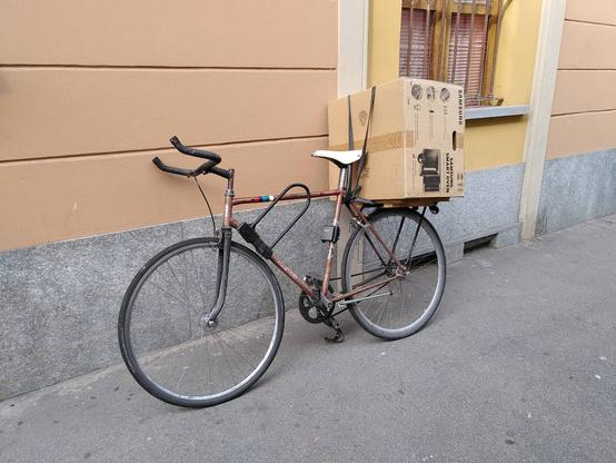 A bicycle is leaning against a house wall with a large box strapped down on the modified rack. The straps are old, cut inner tubes, which works fine unless you tie a knot without a loop - if there's no loop and you tie hard you'll have a fun time undoing the knot later!