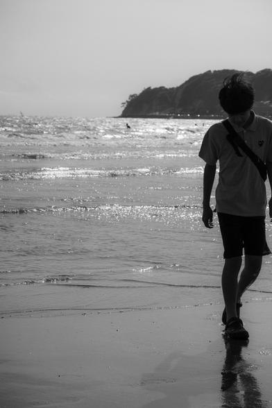 The image is a black and white photo depicting a person walking along a beach. The individual is silhouetted against the background of the ocean, with waves glistening in the sunlight. In the distance, a hilly or cliff-like landform is visible along the coastline.