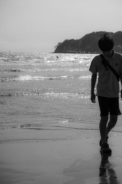 The image is a black and white photo depicting a person walking along a beach. The individual is silhouetted against the background of the ocean, with waves glistening in the sunlight. In the distance, a hilly or cliff-like landform is visible along the coastline.