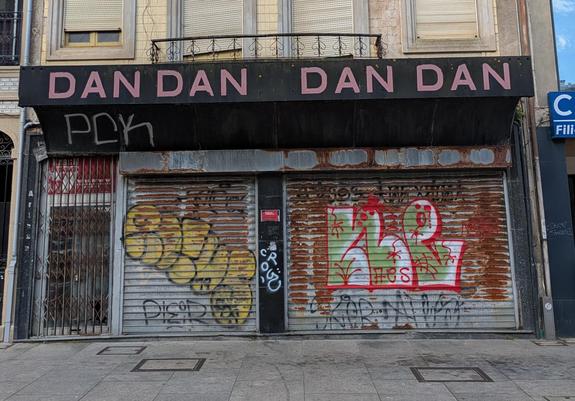 A shop front with a metal gridded gate, closed, to the left and metal shutters pulled fully down over the windows. Theses shutters are liberally graffitied. 
Above is the shop sign, full width of the shop. It is black, and in dark pastel pink sans serif uppercase the shops name - DAN DAN DAN DAN