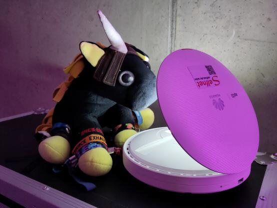 Ida (schwarz-gelbes Flauschehorn) sitzt auf einem Flightcase vor einem Access Point. Der AP ist aufgeklappt, aus seinem innerem kommt Licht. Die Lichtquelle ist nicht sichtbar.