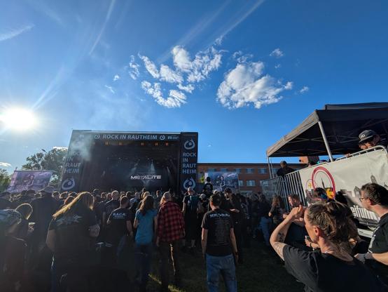 Foto aus der Crowd auf einem Rock/Metalfestival in Richtung der Bühne. Rechts am Bildrand hängt ein Banner von Freifunk Braunschweig, auf der Bühne spielt Metalite. Blauer Himmel mit weniger Wolken, die Sonne steht hinter der Bühne.