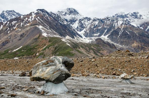 A large, angular boulder sits elevated on a narrow column of white ice on the surface of a glacier. A second, smaller boulder rests similarly nearby. The glacier is covered in patches of gray ice and scattered rocks. Behind the scene, steep, snow-streaked mountains rise under a cloudy summer sky.