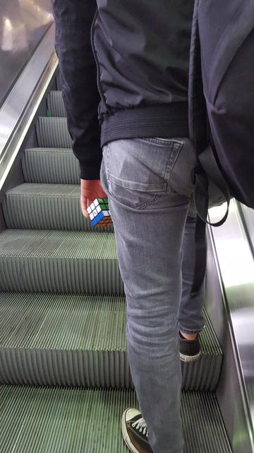 A young person standing on the right side of an escalator going up with the right foot one step higher than the left one. They are seen from the back, visible from just under their shoulders to their feet and are wearing a dark blue jacket, faded blue jeans, low, dark blue, converse like sneakers and a blue bagpack over their right shoulder, their left arm hanging down along their body. 

In their left hand they have a fully solved rubik's cube, apart from the bottom row which is a single turn to the left away from being correct.