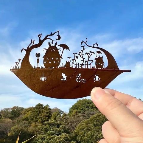 The image features a hand holding a rust-colored metal cutout shaped like a leaf. The cutout depicts a scene from the animated film "My Neighbor Totoro," with characters such as Totoro, a small creature, and a tree with a crescent moon hanging from its branches. The scene includes various elements like mushrooms, plants, and insects, all intricately cut out to create a silhouette effect. The background shows a clear blue sky with some clouds and a lush green landscape, providing a natural contrast to the metal cutout. The hand holding the cutout is visible, with fingers gently gripping the leaf-shaped object.

Provided by @altbot, generated privately and locally using Ovis2-8B