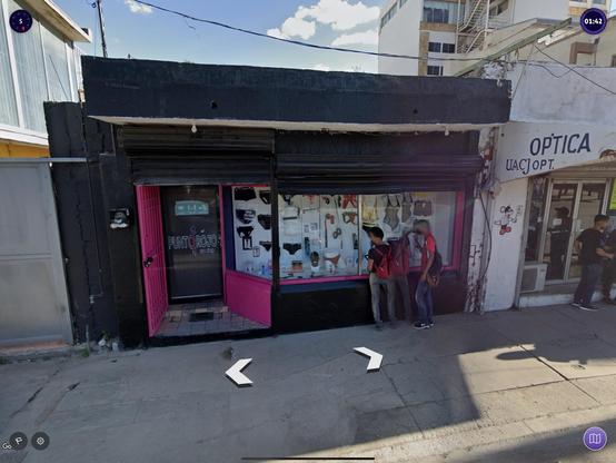 A storefront labeled "Punto Rojo sex shop" features a pink entrance and displays various adult items. Three individuals stand outside, examining the merchandise. Nearby, there is another store labeled "Óptica." The scene is set on a sunny day