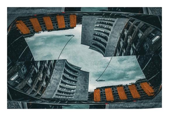 A photo of the top of a car reflecting some buildings. There are two of the same image one on top of the other and mirror opposites. There is a grey brutalist building on the bottom left and the top right while the yellow façade of the other building is on the bottom right and the top left. It is a dark, moody photos with lots of grey and a tumultuous looking sky.