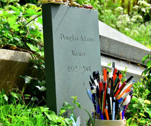 A gravestone inscribed with “Douglas Adams, Writer, 1952–2001” stands amid greenery, with a pot of pens placed in front.

Ein Grabstein mit der Inschrift „Douglas Adams, Schriftsteller, 1952–2001“ steht inmitten von Gruen, davor ist ein Topf mit Stiften aufgestellt.