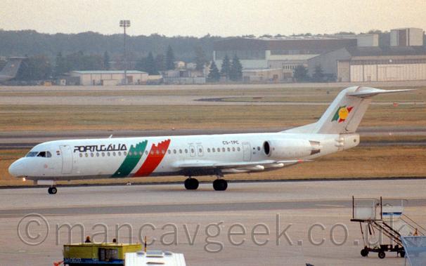 Side view of a white, twin engined jet airliner with the engines mounted on the sides of the rear fuselage, taxiing from right to left.
The plane is mostly white, with diagonal green and red bands running from the top of the body, down and forward under the belly.
There are black "Portugalia" titles on the upper forward fuselage, just aft of the forward door.
The tail is mostly grey, with a smaller version of the green and red bands body bands overlaid with a yellow circle.
Grey concrete apron dills the foreground, with grass and taxiways in the background lead up to cream-coloured hangars and other buildings in the distance, under bright but hazy sky.