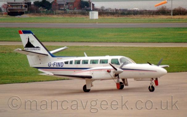 Side view of a white, twin engined BizProp parked facing to the right.
There is a blue and red stripe running along the bidy, as well as over the top section of the engine cowlings over the wings.
The blue registration "G-FIND" is on the upper rear fuselage.
There are black "Air Corbiere" titles on the tail, below a black image of a tower on a domed hill.
Grey concrete apron fills the foreground, with green grass separated by grey taxiways and a black runway leading off into the distance in the background.
A red brick building stands right at the back, with a large body of water just beyond that.
