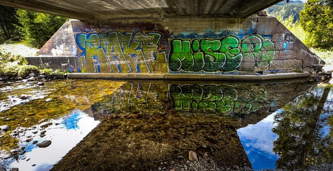Color photo taken under the bridge from the opposite side of the gratitti with concrete "roof" and the river in the foreground. There is bright sun from a partly blue sky, which is reflected in the water. The same is the graffiti on the opposite side.
It consists of a couple of words, which mean nothing to me, in very large and bold letters in different spray colors.

🇸🇯Fargefoto tatt under brua fra motsatt side av grafittien med betong i "taket"  og elva i forgrunnen. Det er strålende sol fra delvis blå himmel, som speiler seg i vannet. Det samme gjør grafittien på motsatt side.
Den består av et par ord, som ikke sier meg noe, i veldig store og uthevede bokstaver i forskjellige sprayfarger.