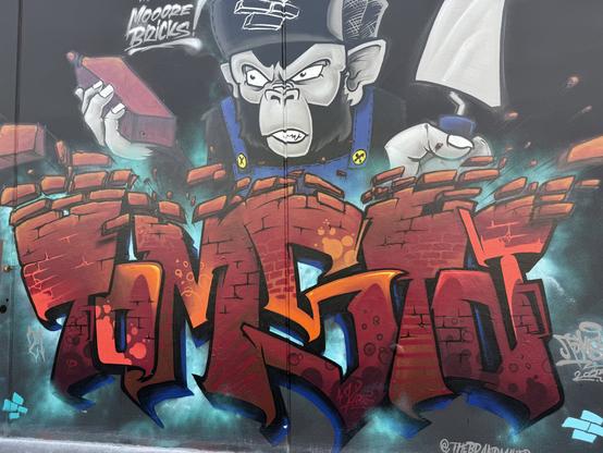 A spraypaint wall art of letters made of brown/red bricks spelling "TOMOSTOT". Above is it an ape holding a brick and a trowel.