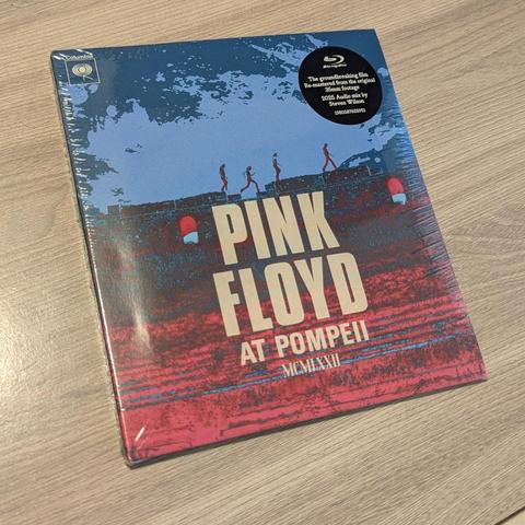 Una imagen de una caja de Blu-ray de Pink Floyd en Pompeya. La caja muestra una imagen de figuras caminando sobre un muro antiguo, con los nombres "PINK FLOYD" y "AT POMPEII" en letras grandes. En la parte superior derecha, hay un pequeño círculo que indica que se trata de un Blu-ray. La fecha "MCMLXXII" (1972) también se muestra en la caja. La caja parece estar nueva y sellada.