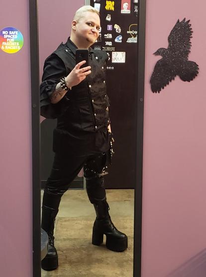 A person in masc goth attire with platform boots taking a mirror selfie at a goth club with a rainbow sticker next to the mirror  that reads "no safe spaces for fascists and racists".  There is a bird silhouette on the other side of the mirror.