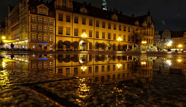 Das Bild zeigt eine nächtliche Szene einer Stadt mit historischen Gebäuden, die sich in einer Wasserfläche spiegeln. Die Gebäude sind beleuchtet, und die Lichter reflektieren sich ebenfalls im Wasser. Links im Bild ist ein Café mit Menschen zu sehen, die draußen sitzen. Die Architektur der Gebäude ist detailliert und zeigt viele Fenster und Verzierungen.