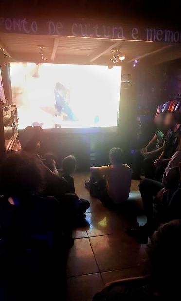 Foto de diversas pessoas sentadas em cadeiras e no chão assistindo um filme em um telão em um ambiente escuro.