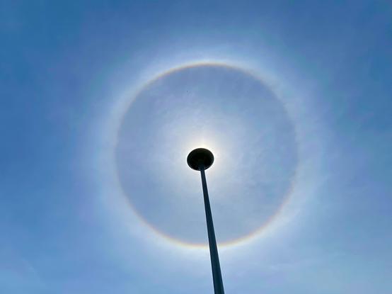 The top of a lamppost is blocking the sun, around that silhouette is a colourful circular halo in a hazy blue sky.