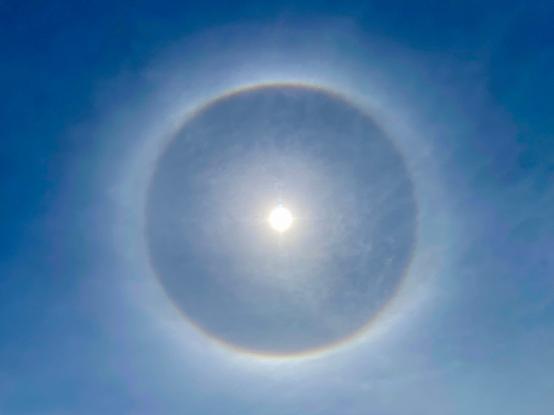 The halo is photographed without anything blocking the sun. The colourful circle appears to have pale bulges to the left and right.