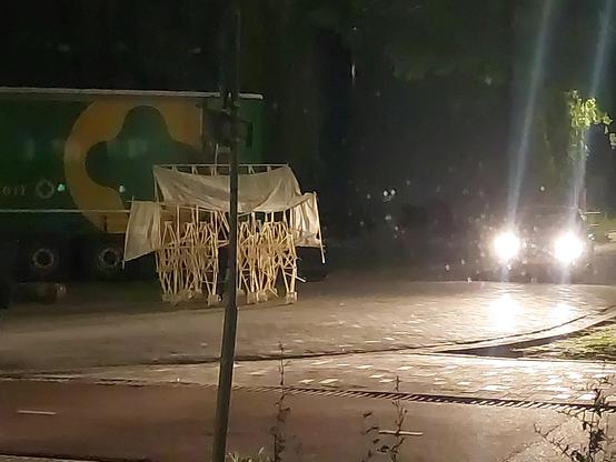 An image of a strandbeest (a sort of walking construction construcred out of pvc pipes), at night, crossing the street (I think a person was walking it). The headlights of a car are visible, there is also a parket truck and a bike lane in the picture