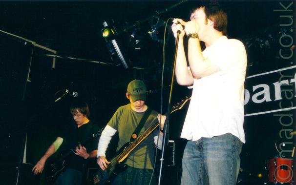 3 members of a band performing on a stage.
On the left is a man with mid-length hair, wearing a black shirt and dark blue jeans, playing a black electric guitar, looking down towards his instrument.
In the middle is a man wearing a khaki green hat, a green t-shirt over a white jumper, and a blue pair of jeans, playing a black bass guitar.
On the right is a man with short brown hair, wearing a white shirt and a blue pair of jeans, holding on to a mic stand as if his life depends on it.
Black walls and ceiling fill the rest of the frame, with large white letters "AR" on the rear wall, part of the word "Barfly" on the wall on the right of the frame.