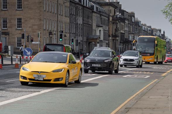A city street scene in Edinburgh, Scotland, showing a yellow Tesla taxi with the plate "TAXI," followed by a black car, a white car, and a yellow-green coach bus, with historic stone buildings and traffic lights in the background.