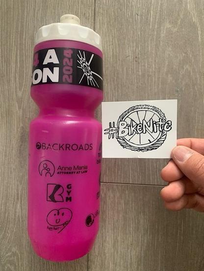 White #BikeNite sticker, black hand drawn text with a wheel in background. Pink cycling water bottle next to for scale.