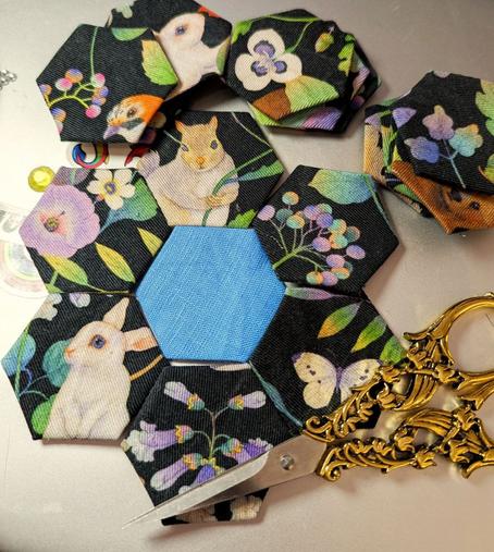 Gray background, on this is a hexagon patchwork flower with an indigo blue center piece laid out. The six "petals" are made from black cloth with a colourful floral pattern and cute, small animals (bunnies, butterfly...). Thee additional staples with basted hexagons are lying alongside. There are also sewing scissors to be seen. The handle is highly ornamented and golden. It looks like a pattern of lily of the valley.