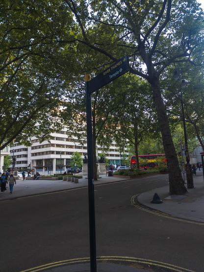 View past a fngerpost direction sign saying 'River Thames' and 'Temple' (latter with a London Underground icon). Behind it is a wide road, then an open area with seats for the public, and buildings going down a hill.