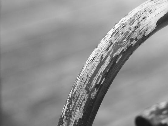 Black and white photo of a curved wicker chair arm, the worn paint revealing the textured wood underneath. The background is softly blurred.