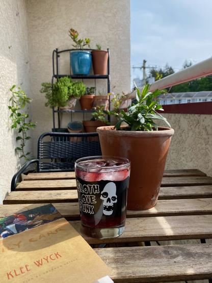 View along a long, narrow balcony. In the foreground is a table with a copy of Emily Carr's book Klee Wyck, a highball glass containing sangria with a skull and the words 'Sloth love punk' painted on it, and a tarragon plant in a pot. In the background is a patio chair, a jasmine growing up a wall, and a shelf with various herbs in pots. A honeysuckle is wrapped around the railing on the right.