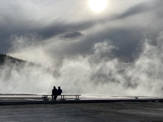 Two people are sitting close to each other on a bench, they are silhouettes in front of sunlit steam coming from in front of them from the Grand Prismatic Spring in Yellowstone

The image looks black and white although it is in color, the sky is partly cloudy and melds with the rising steam which is brighter than the sky