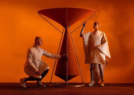 Two figures in stylish, textured outfits engage with a large, funnel-shaped object against an orange backdrop, creating a dynamic scene.