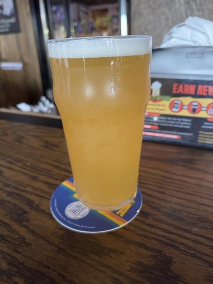 A pint glass filled with a pale, cloudy beverage sits on a circular coaster on a wooden table. The drink has a foamy head and appears to be a type of beer. In the background, bar elements are visible, including a sign.
