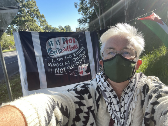 Masked woman stands in front of a “it’s not controversial to say that killing children is not self defence” sign at Bega