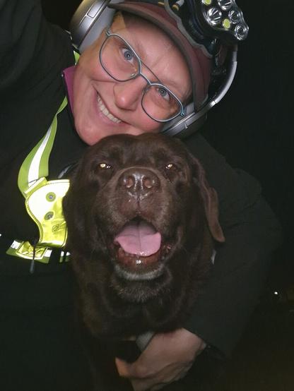 Night time selfie of a person and a chocolate brown Labrador with greying snout. The person is wearing a black jacket, yellow safety harness, red cap, silver headphones and blue-rimmed glasses. On their cap rests a headstrap torch in off position. Their free hand is around the dog's shoulders. Both are facing the camera. The dog's mouth is slightly open, showing off some blep. The person's head is resting on the dog's head. They're smiling, grateful for their time spent together.