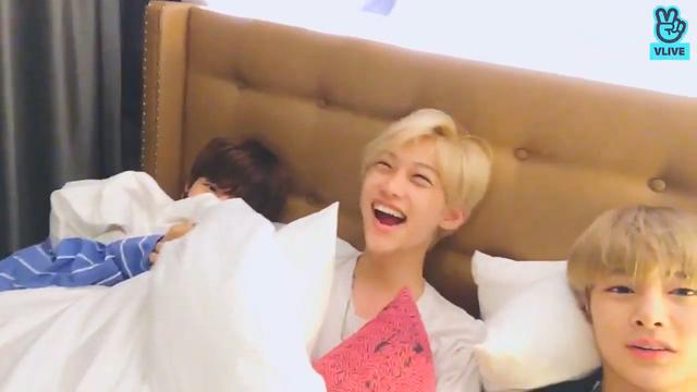 Seungmin, Felix and I.N in a hotel room bed together. Seungmin's peeking out from under the duvet, Felix is laughing with his head thrown back, and I.N is looking at the camera.