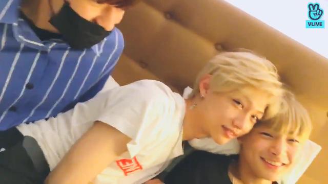 Seungmin, Felix and I.N on I.N's bed. Felix and I.N have their heads close together, and are smiling at the camera. Seungmin is behind Felix, is wearing a face mask, and has a slightly manic air.