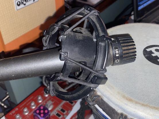 The image shows a microphone mounted in a shock mount, positioned above a percussion instrument. The background includes an amplifier and other music equipment.