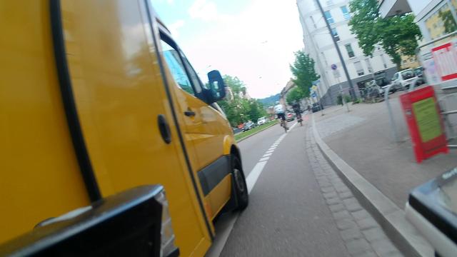 POV Radfahrer: ein völlig unterdimensionierter Radfahrstreifen in einer leichten Rechtskurve. Links zu sehen ist ein DHL-Fahrzeug, das mit Vorderreifen, Spiegel und Aufbau rechts der Trennlinie fährt.