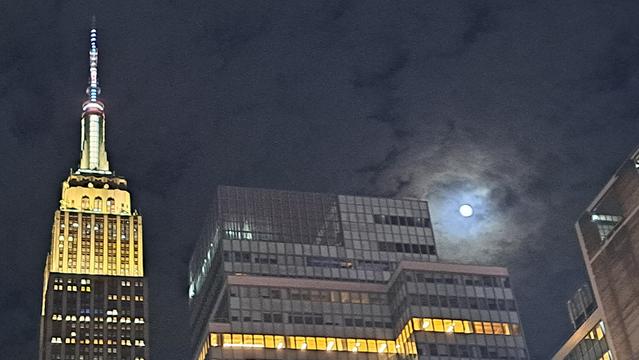 Night shot showing the top of Empire State Building on the left while on the right the moon peeps through the clouds